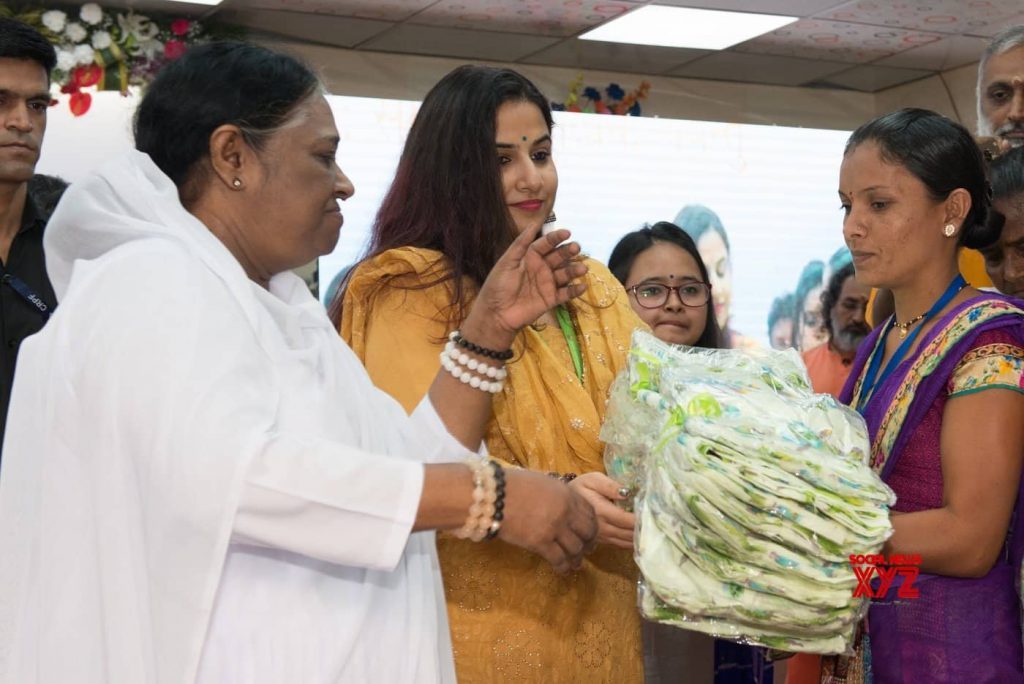 Amma in the foreground and Vidya Balan next to her in yellow hand a bundle of light green saris to a woman in purple. 