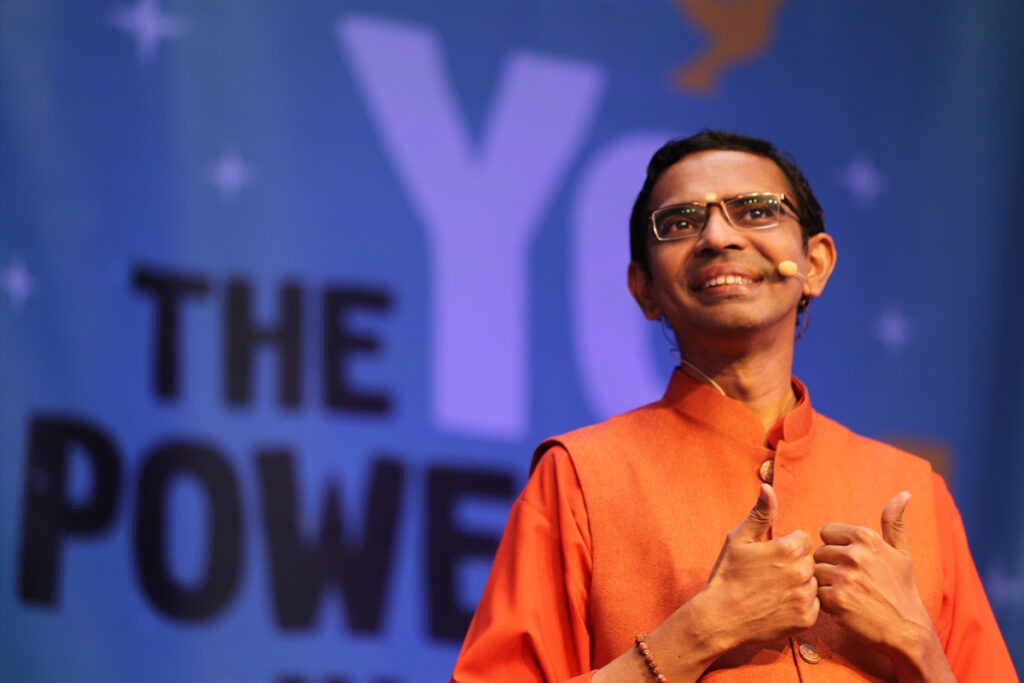 Swami Shubamritananda gives two thumbs up, smiling, as he stands on the stage.