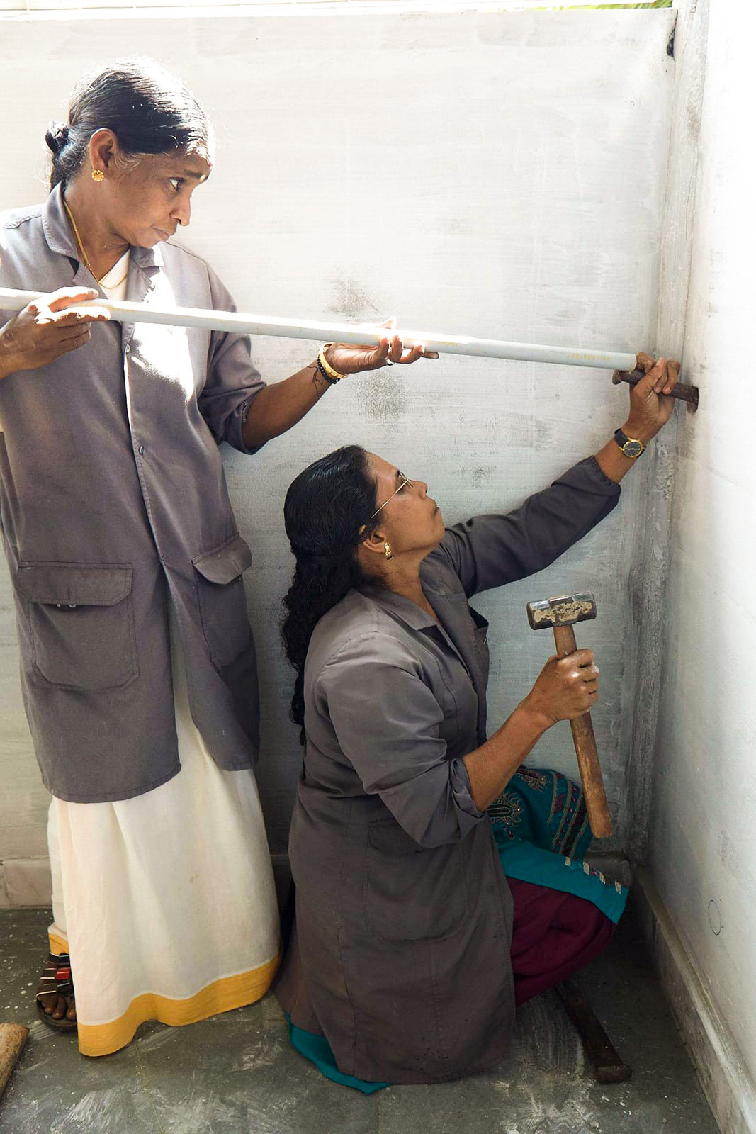 Two village women wearing dark grey work coats, concentrate as they insert a plumbing pipe into the wall.