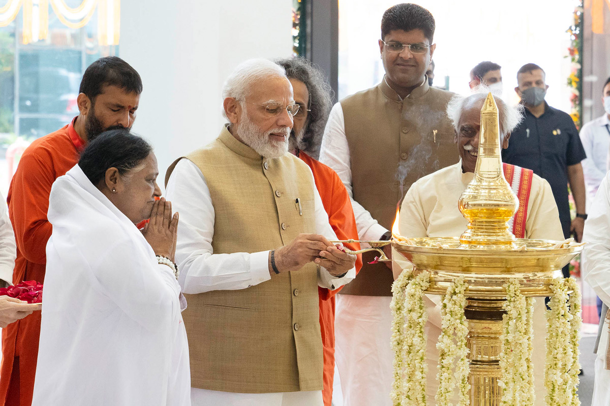 Amma, Swami Amritaswarupananda, Swami Nijamrita and others watching with hands in prayer as PM Modiji lights a large lamp as a traditional form of worship.