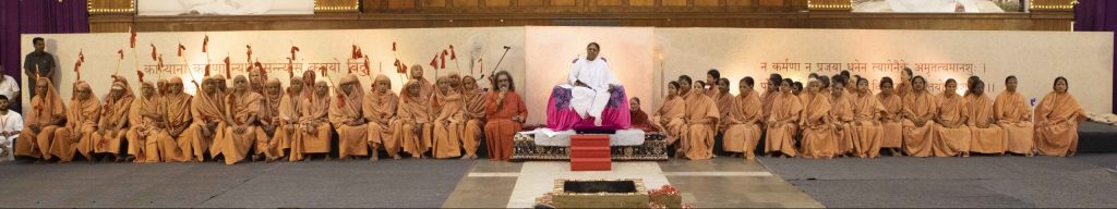 Amma is seated center on a pink and purple peetham, and Swami Amritaswarupananda sits to her right speaking to the male and female sannyasi initiates.