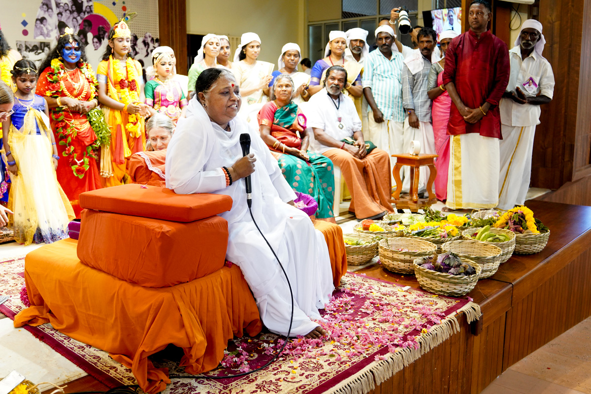 Amma sits on her peetham with the mic surrounded by Amrita Niketanam alumni dressed in colorful dance costumes, saris and smiles. Chief Guests – Nanjiyamma, renowned Irula folk singer, and Cheruvayal Raman from Wayanad who won the Padma Shri award for his efforts to conserve indigenous rice varieties stand nearby.